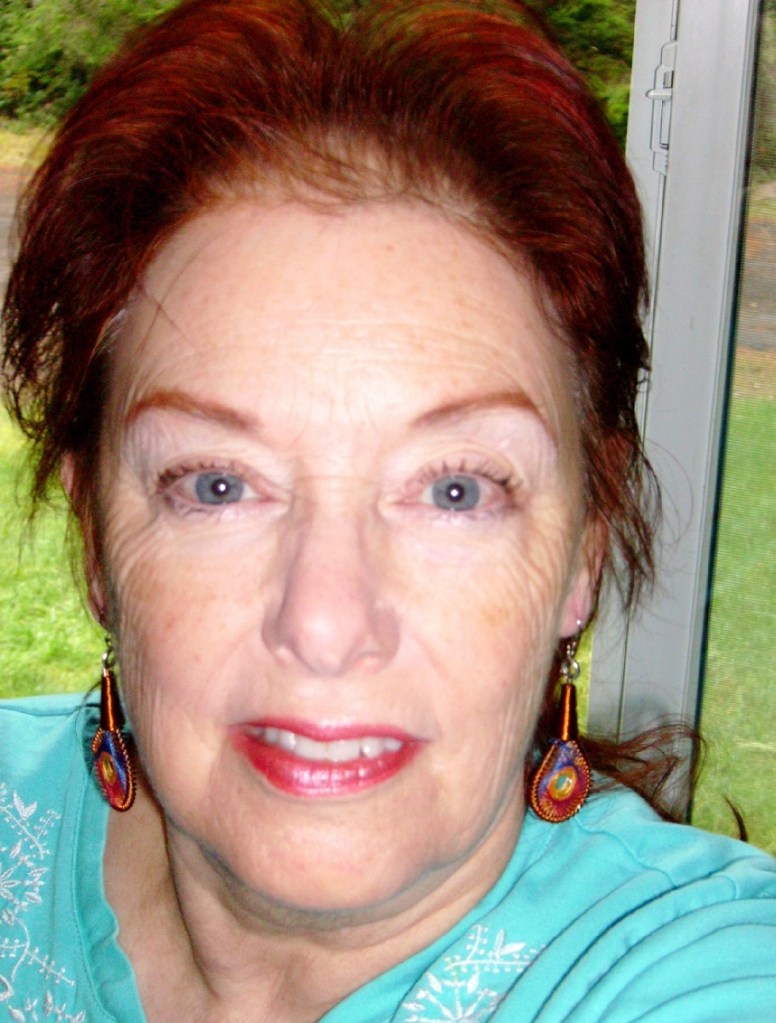 Dana Corby has red hair and blue eyes. She is indoors by a window looking out to a garden. Her top is a light aqua. Her earrings are long and red and blue.