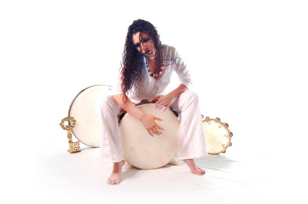 Arlechina has long dark, curly hair. She is sitting on top of a large hand drum dressed in white with one hand on the drum head. Behind her are another large hand drum and a smaller hand drum with a tambourine edge. leaning against the large drum in the background is a gold-colored key that is about the same length as Arlechina's  forearm.